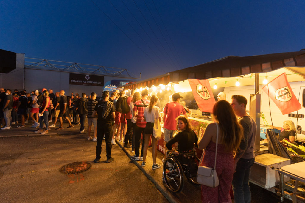 Dss membres du public font la queue devant les stands de sandwichs et, en arrière-plan, pour accéder au stade. Après une année de matches à huis-clos en raison de la crise sanitaire, le public a pu de nouveau se rendre au stade Ernest Wallon à l’occasion du match de clôture de la 2e journée du Top 14. Champion de France en titre, le Stade Toulousain recevait le Rugby Club Toulonnais. Le match s'est joué à guichets fermés et près de 19000 contrôles de pass sanitaires ont été effectués à l'entrée du stade. Le Stade Toulousain l’a remporté par 41 à 10. Toulouse, stade Ernest Wallon, le 12 septembre 2021.