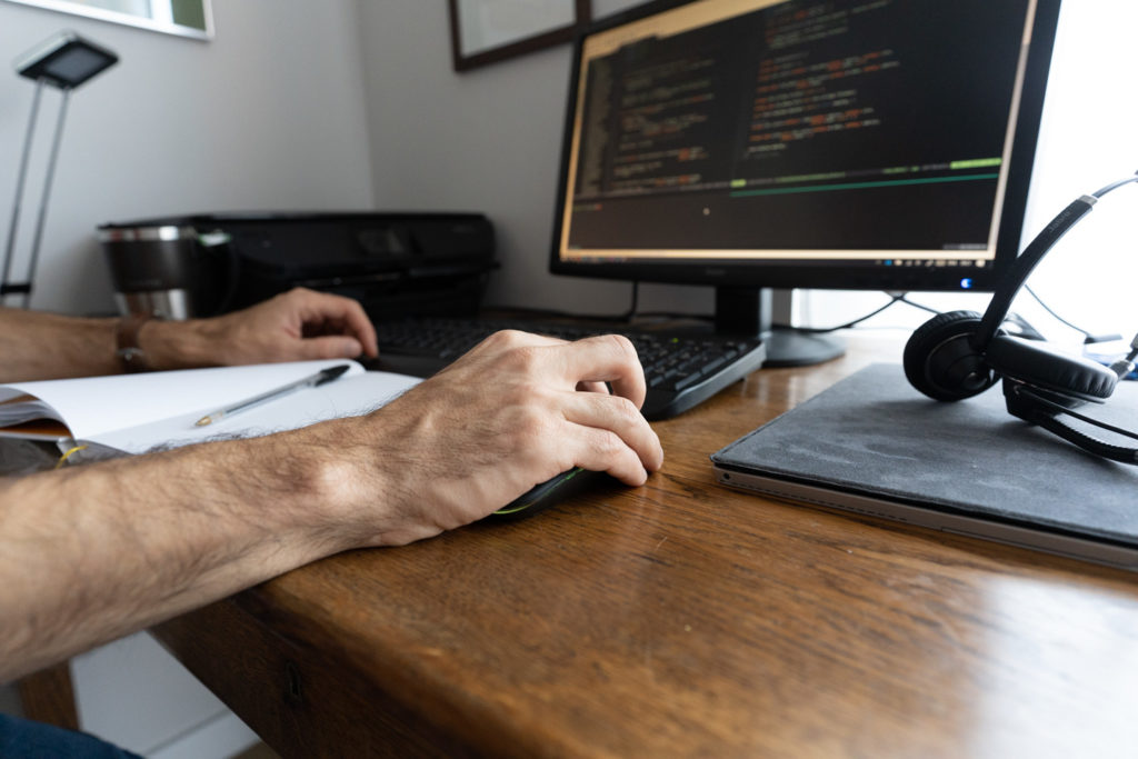 Poste de travail - avec écran, clavier, casque et cahier - d'un informaticien chez lui en télétravail. Sa main droite est posée sur la souris.