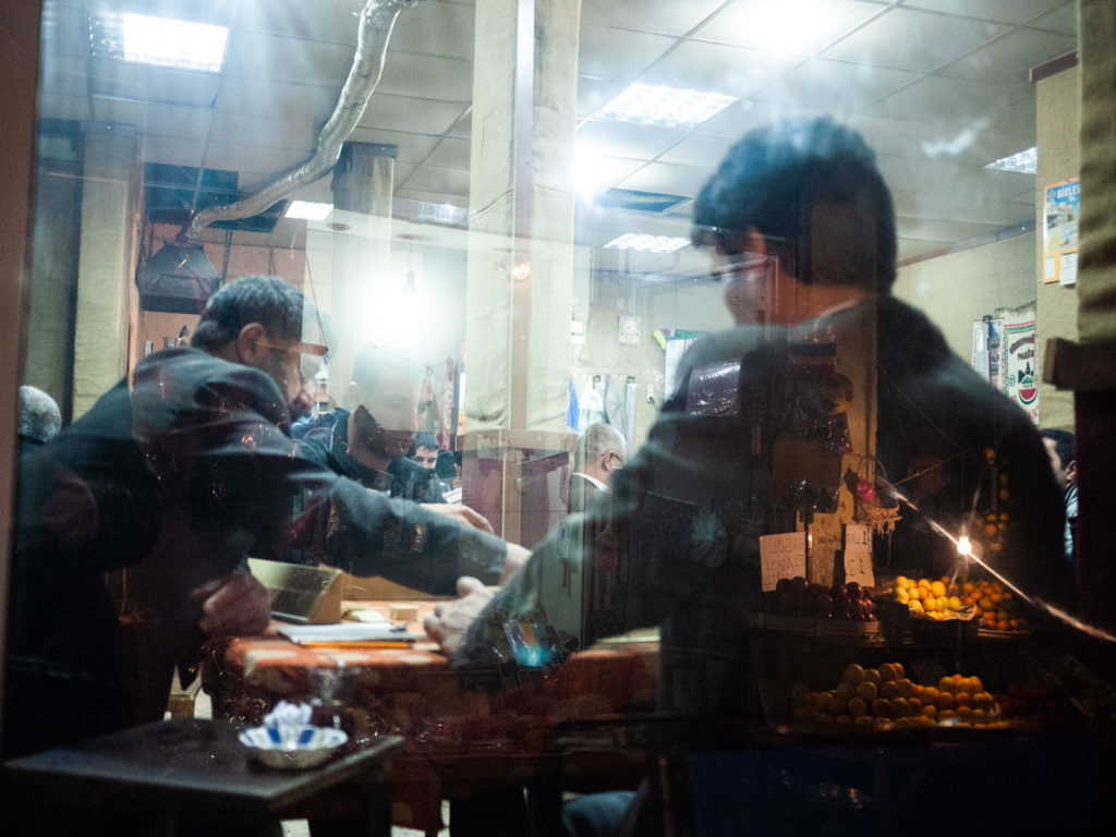 Un groupe d'hommes jouent à "Okey" - un jeu de dominos - dans une maison de thé traditionnelle uniquement fréquentée par des hommes. En reflet dans la vitrine, un l'étal d'un marchand de légumes. Diyarbakir (Turquie), novembre 2010.